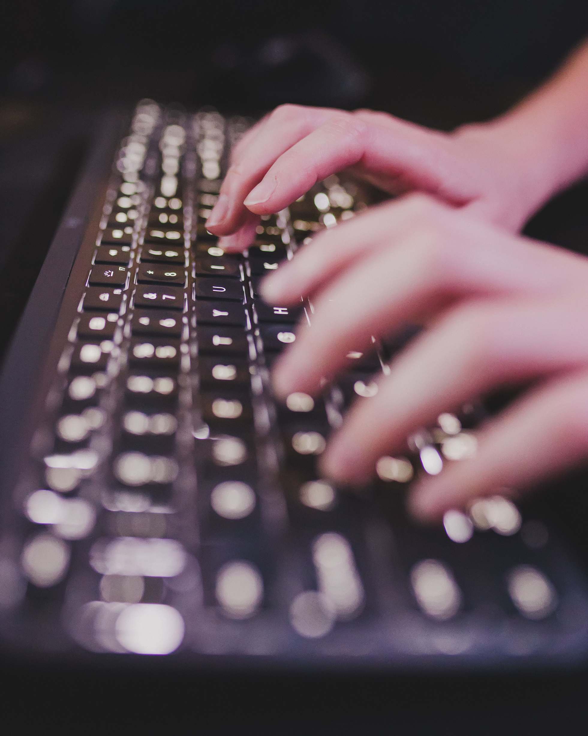 Hands on a laptop keyboard in a workspace typing on an illuminated keyboard